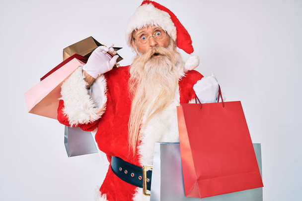 Old senior man with grey hair and long beard wearing santa claus costume holding shopping bags in shock face, looking skeptical and sarcastic, surprised with open mouth  - Fotó, kép