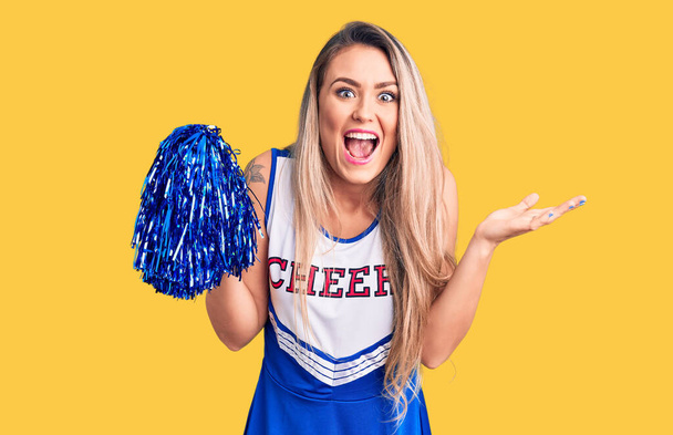 Young beautiful blonde woman wearing cheerleader uniform holding pompom celebrating victory with happy smile and winner expression with raised hands  - Photo, Image
