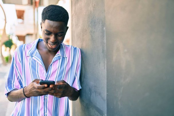 Jovem afro-americano sorrindo feliz usando smartphone apoiado na parede na rua da cidade. - Foto, Imagem