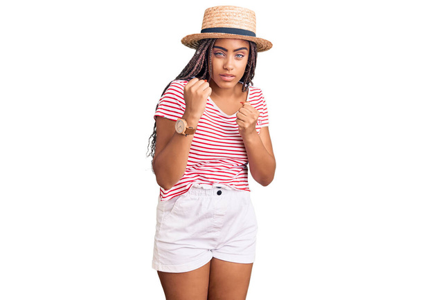 夏の帽子をかぶっている若いアフリカ系アメリカ人女性は、拳の防衛ジェスチャー、怒りと動揺した顔、問題を恐れて戦う準備ができています  - 写真・画像