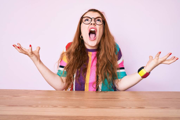 Young redhead woman sitting on the table wearing glasses crazy and mad shouting and yelling with aggressive expression and arms raised. frustration concept.  - Photo, Image