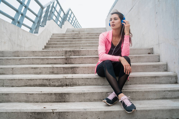 Retrato de una mujer atlética escuchando música en un descanso del entrenamiento mientras está sentada en las escaleras. Concepto de estilo de vida deportivo y sanitario. - Foto, imagen
