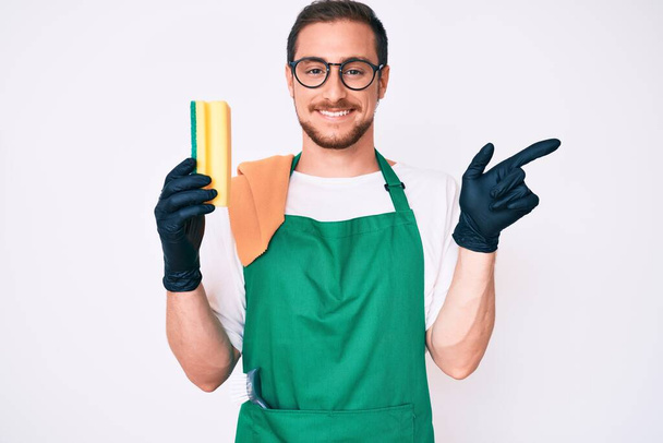 Young handsome man wearing apron holding scourer smiling happy pointing with hand and finger to the side  - Photo, Image