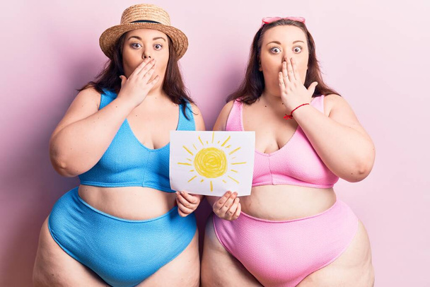 I giovani gemelli plus size che indossano bikini che tengono il sole disegnano la bocca di copertura con la mano, scioccati e spaventati per errore. espressione sorpresa  - Foto, immagini
