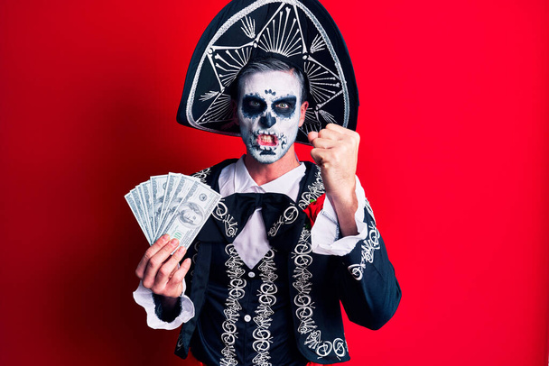 Jongeman draagt Mexicaanse dag van het dode kostuum met dollars in zijn hand geïrriteerd en gefrustreerd schreeuwend van woede, gek schreeuwend van woede en met de hand opgestoken  - Foto, afbeelding
