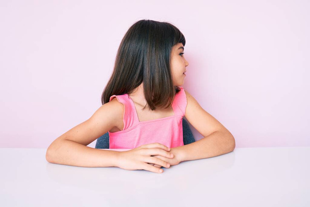 Fiatal lány bumm visel alkalmi ruhák ül az asztalon keres oldalra, pihenjen profil pózol természetes arc magabiztos mosollyal.  - Fotó, kép