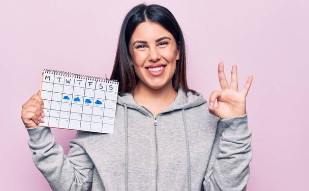 Young beautiful brunette woman holding weather calendar showing rainy week doing ok sign with fingers, smiling friendly gesturing excellent symbol - Fotó, kép