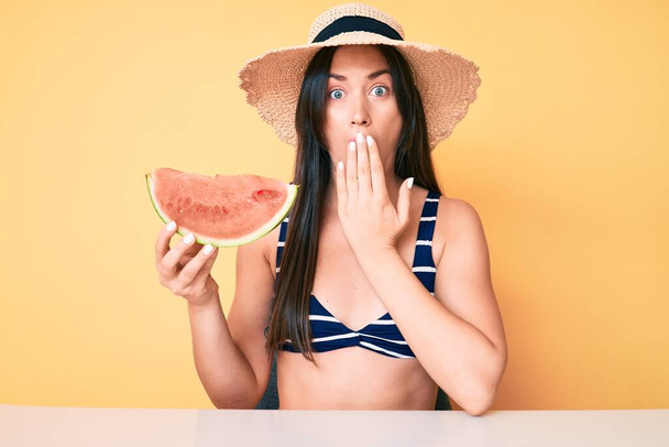 Jeune belle femme caucasienne portant un bikini et un chapeau tenant une tranche de pastèque couvrant la bouche avec la main, choquée et effrayée par l'erreur. expression surprise  - Photo, image