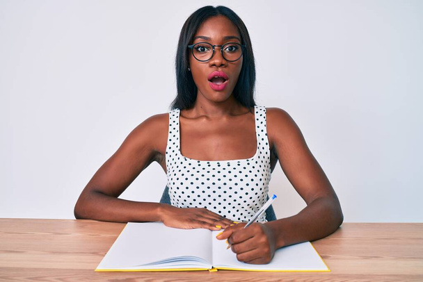 Młoda Afroamerykanka pisze książkę siedząc na stole przerażona i zdumiona otwartymi ustami dla zaskoczenia, niedowierzania twarz  - Zdjęcie, obraz