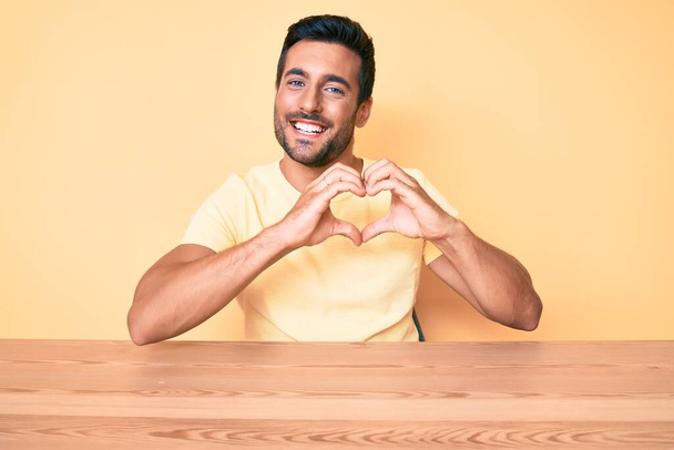 Junger hispanischer Mann in lässiger Kleidung sitzt auf dem Tisch und lächelt verliebt und zeigt Herzsymbol und Form mit den Händen. Romantisches Konzept.  - Foto, Bild