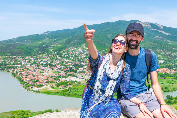 Una pareja emocionada y amorosa se sienta en el lado derecho y señala hacia arriba con el fondo de montañas verdes y la ciudad histórica de Mtskheta. Viajes y tours en Georgia. - Foto, imagen