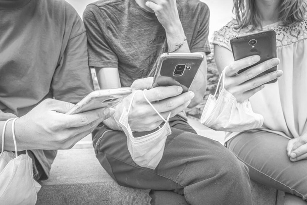 Černobílý zblízka okamžik tři osoby v neformálních barevných šatech sedí a prohlížejí si telefony s maskami v rukou. Sociální média a interagující životní styl během pandemie 2020 - Fotografie, Obrázek