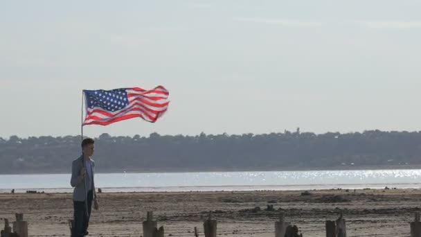 Ένας πατριώτης των ΗΠΑ με τη σημαία στα χέρια του βόλτες κατά μήκος του ποταμού παραλία, δείχνοντας το χέρι του προς τα εμπρός. - Πλάνα, βίντεο