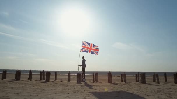 Um patriota do Reino Unido monta um giroscópio ao longo de postes de madeira com uma bandeira na mão contra um céu azul. - Filmagem, Vídeo