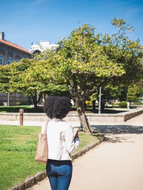 Vista trasera de una joven negra, con el pelo rizado, sosteniendo algunos libros, afuera - Foto, imagen