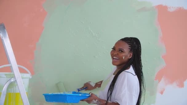 Concepto de renovación plana. Mujer afroamericana sonriente redecorando su casa sosteniendo un rodillo de pintura cubierto de pintura naranja y verde - Metraje, vídeo