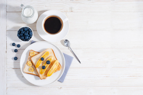 Breakfast with bananas and blueberry with cup of coffe and baked toasts on the white wooden table flat lay with copy space. Healthy tasty food at the morning with oat-flakes - Photo, image