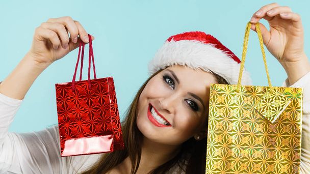 Young brunette woman is happy to give Christmas gifts. Female wearing santa claus hat holding presents festive gift bags, on blue - Fotó, kép