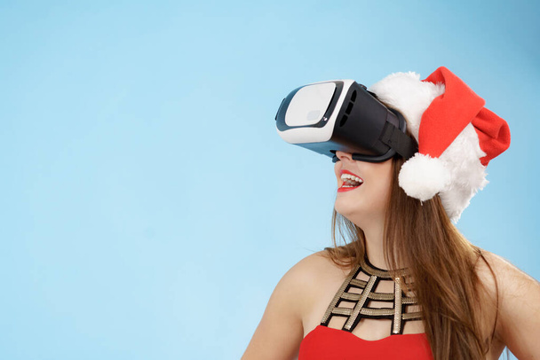 Woman wearing santa claus hat exploring space with virtual reality goggles headset. Amazed girl watching 3d film tour in vr glasses box. New generation cyber christmas concept, on blue. - Foto, imagen