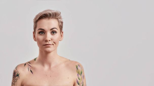 Well... Portrait of a young attractive half naked tattooed woman with short hair looking with smirk at camera being disappointed or unimpressed isolated over light background - Foto, afbeelding
