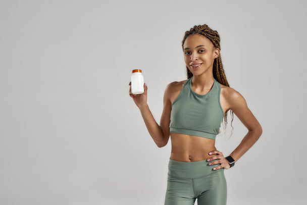 Sport, healthcare and diet concept. Young athletic slim mixed race woman in sportswear holding bottle with capsules or pills and looking at camera - Фото, зображення
