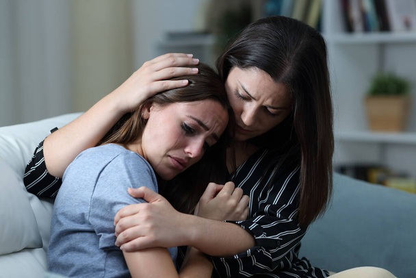 Sad teen crying being comforted by her sister on a couch in the living room at home  - Foto, Bild