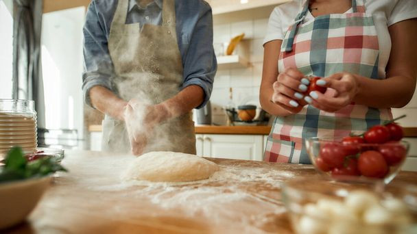 Cropped shot of couple preparing for making pizza together. Young man making the dough while woman in apron preparing tomatoes. Hobby, lifestyle - Foto, Imagen