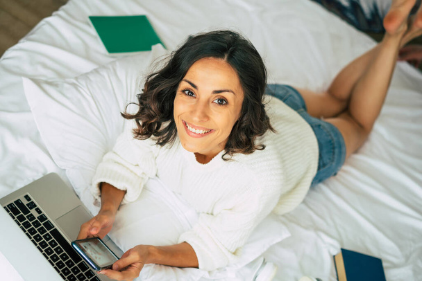 Happy beautiful young brunette woman is working with her laptop while she lay in bed at home. The freelance girl is surfing on internet - Φωτογραφία, εικόνα