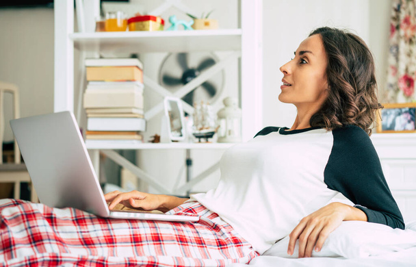 Happy beautiful young brunette woman is working with her laptop while she lay in bed at home. The freelance girl is surfing on internet - Фото, зображення