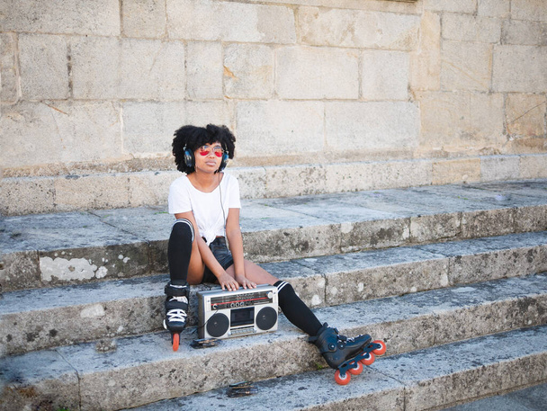 Joven mujer negra sentada en unas escaleras en la calle, escuchando música con auriculares y una radio de cassette vintage - Foto, Imagen