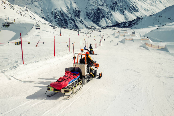Снегоходная машина с санями и снаряжением быстро поднимается на водителя, чтобы помочь травмированному лыжнику или сноубордисту при несчастном случае на зимнем горном курорте. Спасательная машина на горнолыжном спуске - Фото, изображение