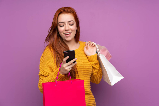Redhead teenager girl over isolated purple background holding shopping bags and writing a message with her cell phone to a friend - Фото, изображение