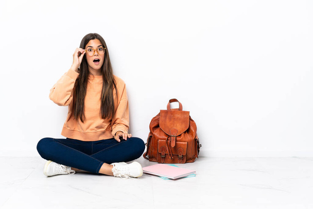 Young student brazilian woman sitting on the floor with glasses and surprised - Photo, Image