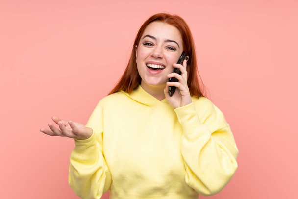 Adolescente rousse sur fond rose isolé en gardant une conversation avec le téléphone mobile avec quelqu'un - Photo, image