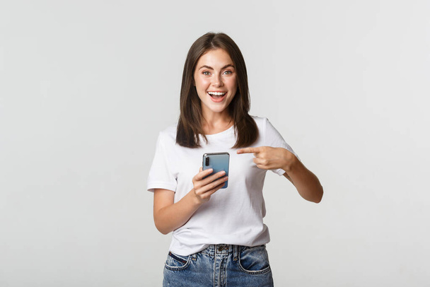 Excited and pleased brunette girl pointing finger at smartphone screen - Φωτογραφία, εικόνα