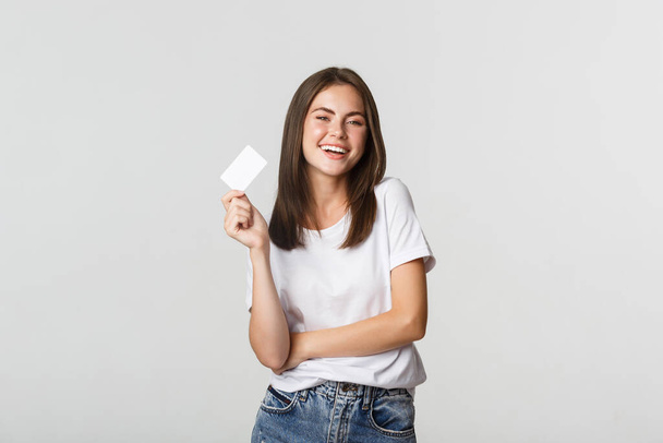Ελκυστική ευτυχισμένη μελαχρινή κοπέλα γελώντας και κρατώντας πιστωτική κάρτα, λευκό φόντο - Φωτογραφία, εικόνα