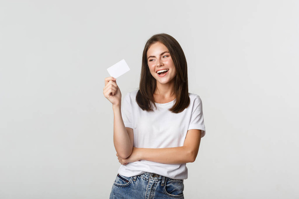 Aantrekkelijk gelukkig brunette meisje lachen en houden credit card, witte achtergrond - Foto, afbeelding