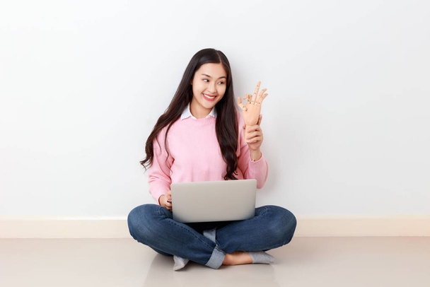 Fiatal vonzó ázsiai nő, mint egy diák gazdaság fa kéz mutatja ok gesztus laptop a lábán, online oktatási koncepció. - Fotó, kép