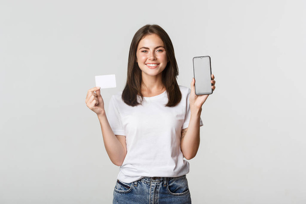 Attraente ragazza sorridente che sembra soddisfatta e mostra carta di credito, schermo del telefono cellulare - Foto, immagini
