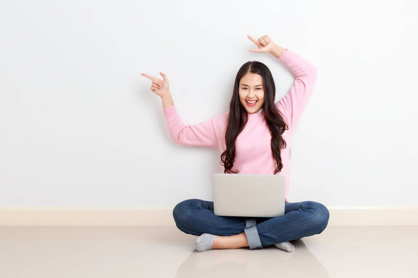 Young attractive Asian woman sitting with laptop on her legs finger pointing up showing copy space with smiley face. - Fotoğraf, Görsel