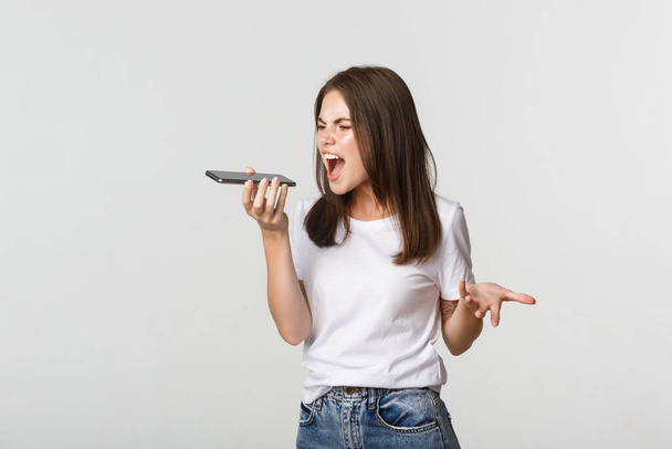 Angry, pissed-off brunette girl arguing, record voice message on mobile phone, white background - Foto, immagini