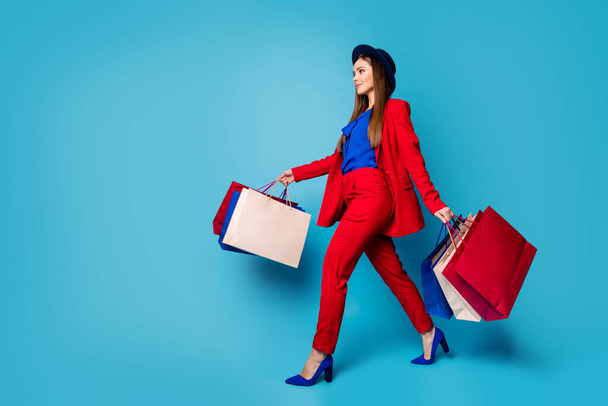 Full body profile photo of self-confident business lady boss walking shopping center holding many packs wear retro cap red suit shirt blazer pants high-heels isolated blue color background - Zdjęcie, obraz