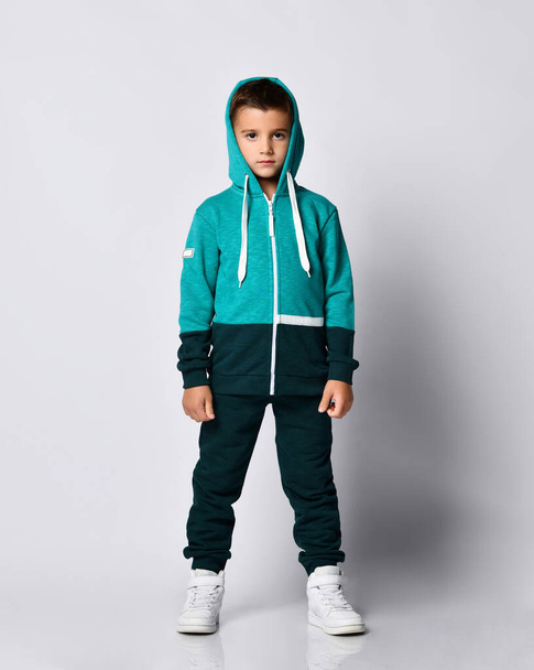 Little brunette kid in colorful tracksuit and sneakers on a gray background. - Foto, immagini