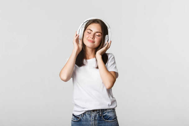 Мечтательная красивая девушка наслаждается прослушиванием музыки в беспроводных наушниках, улыбаясь счастливой - Фото, изображение
