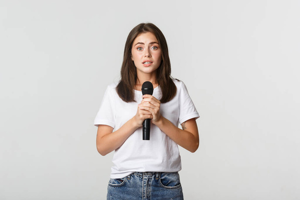 Shy cute brunette girl scared of singing in public, standing with microphone and looking nervous - Photo, Image