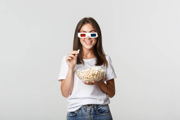 Üç boyutlu gözlüklü, film ya da televizyon dizisi izleyen, patlamış mısır yiyen ve gülümseyen neşeli genç bir kadın. - Fotoğraf, Görsel