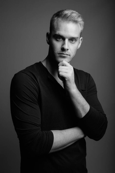 Studio záběr mladého pohledného muže s blond vlasy na sobě dlouhé rukávy košile proti šedé pozadí v černé a bílé - Fotografie, Obrázek