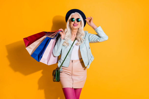 Portrait of positive cheerful old woman have leisure travel trip walk enjoy shop hold bags wear good look leather skirt glasses headwear isolated over bright shine color background - 写真・画像