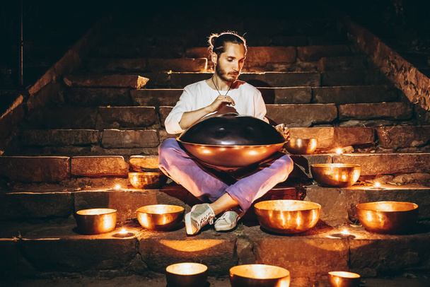 silhouette of a man playing handpan surrounded by Tibetan singing bowls and lighted candles sitting on the steps of the night city. Noise effect on the film - Foto, immagini