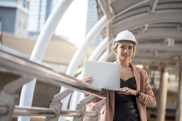 Gyönyörű amerikai építész mérnök nő kemény kalap és kabát ellenőrizze a tervrajzot a laptopon, és nézd meg az építési telek a városban. Vonzó munkavállaló ellenőrizze a projekt terv vagy licitárverés interneten. - Fotó, kép